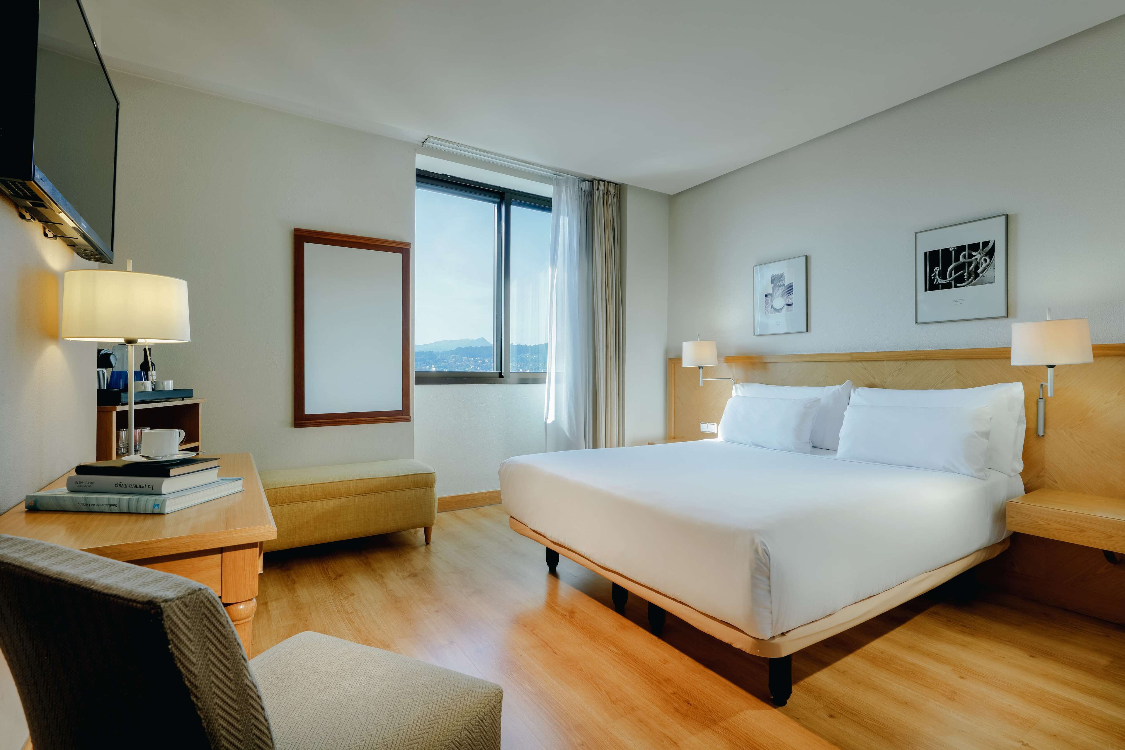 Una habitación de hotel con una cama grande, un escritorio con lámpara, una silla y un ventanal con cortinas.
