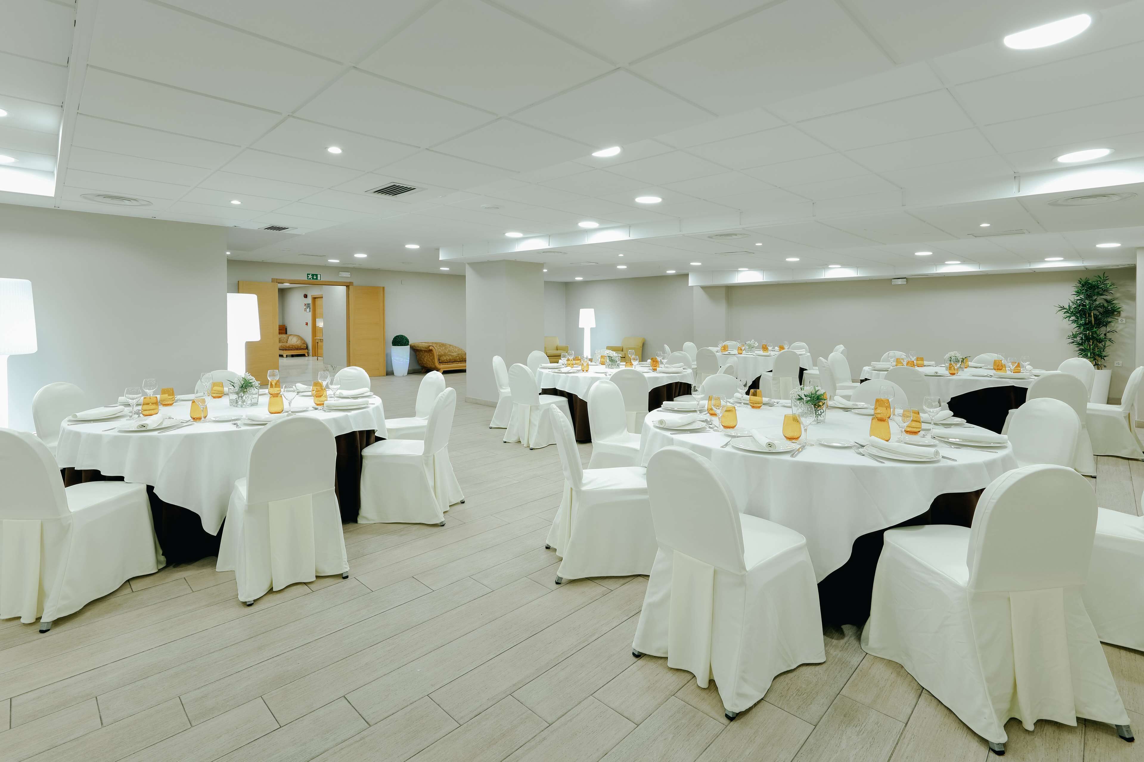 A large conference room with multiple round tables, white chairs, and tables set with orange napkins and glassware.