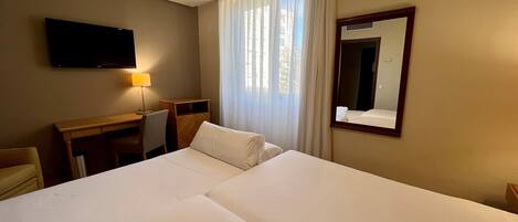 Habitación de hotel con cama, escritorio, silla, lámpara, televisor y ventana con cortinas.