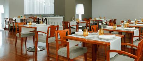 Un comedor con mesas y sillas de madera, manteles blancos y un amplio ventanal que proporciona luz natural.