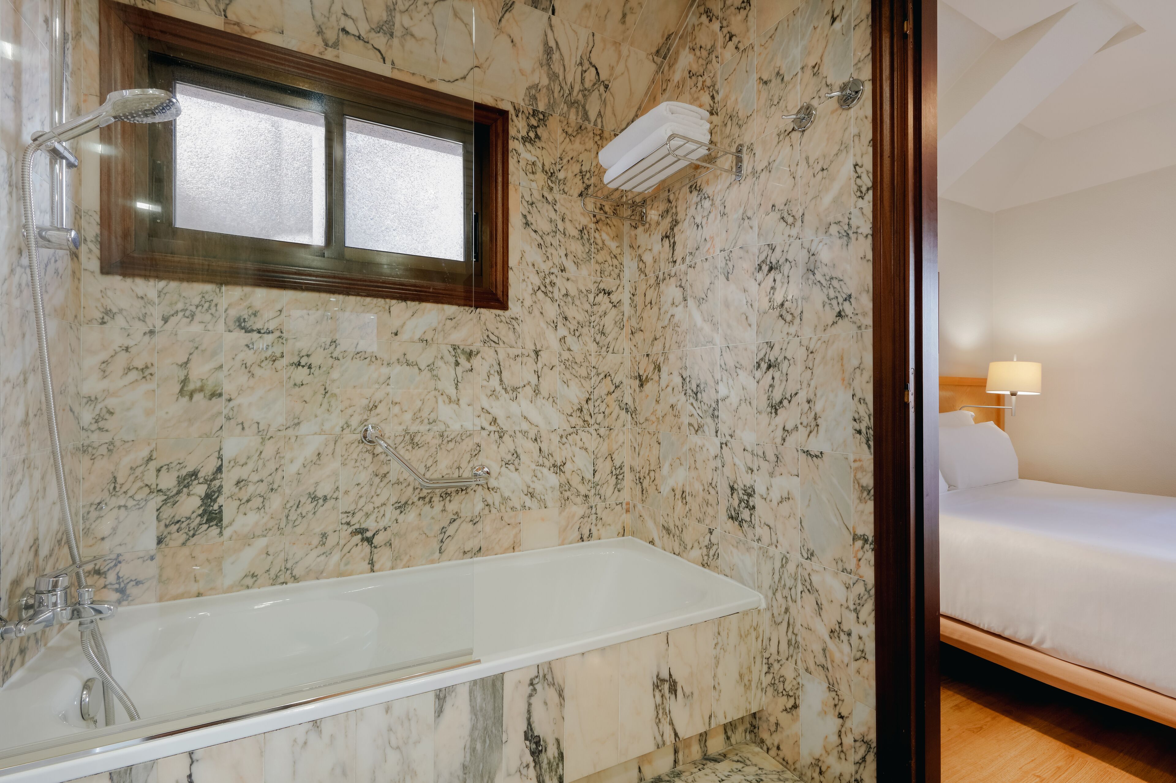 Un baño con pared de mármol, bañera, ducha y una cama con mesita de noche.