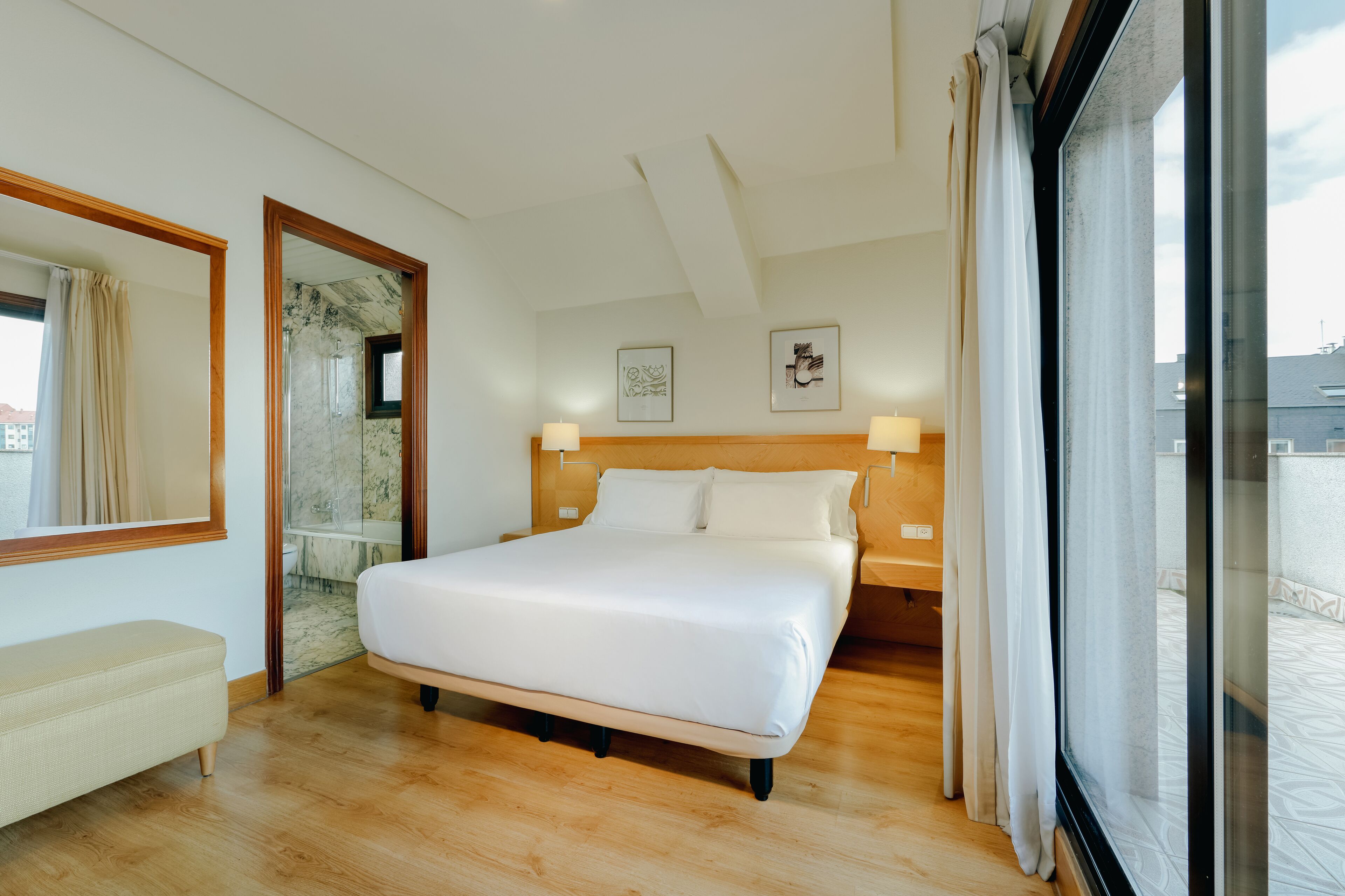 A hotel room with a large bed, a bench, a wooden headboard, and a view through a glass door.