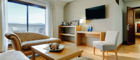 A hotel room with a sofa, armchair, and a small table with decorative items. There is a TV mounted on the wall and a door leading to another room.