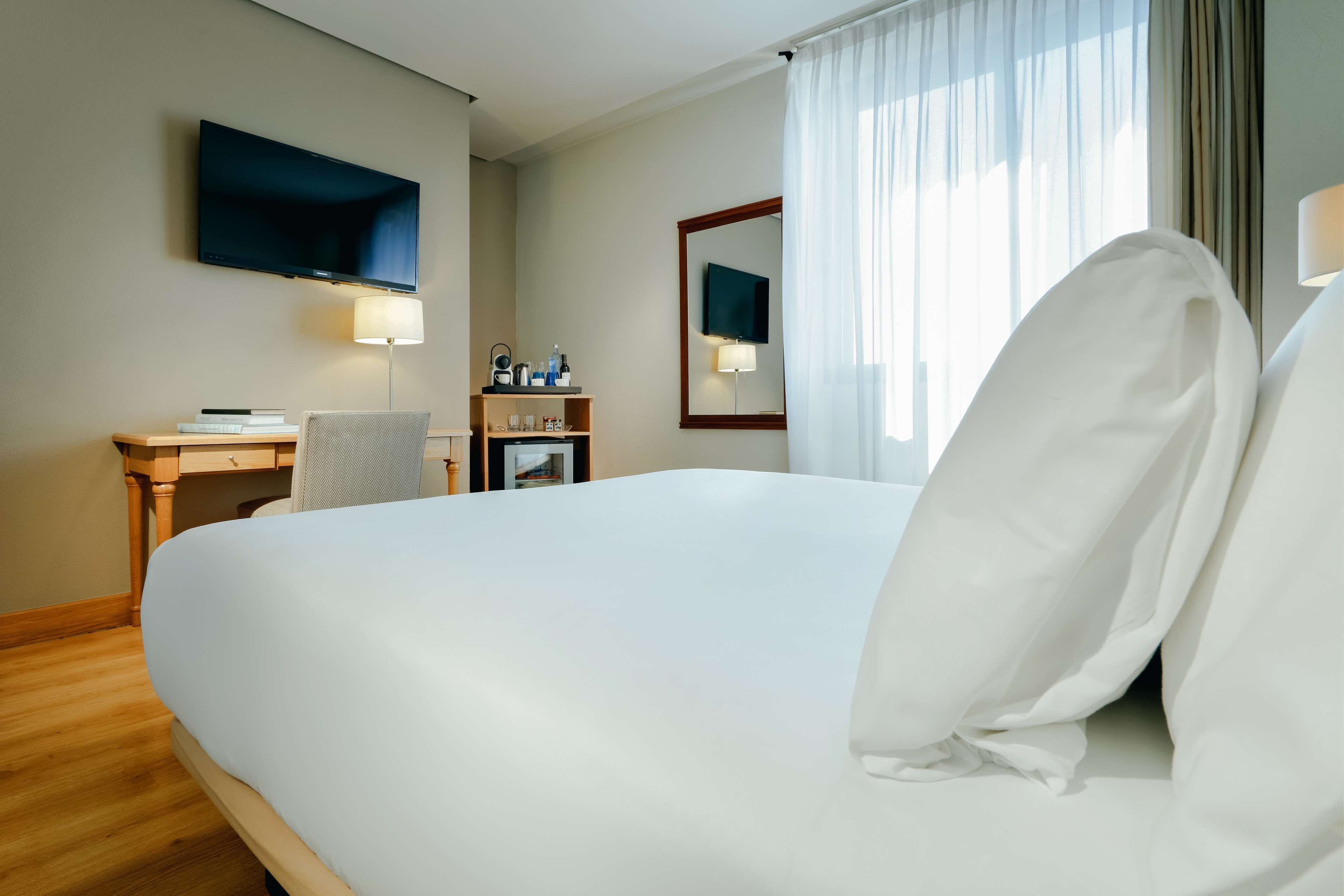 A hotel room with a large bed, a desk with a chair, a TV mounted on the wall, and a window with curtains.