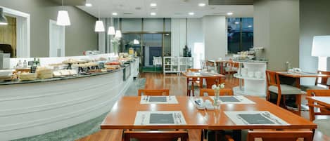 Un comedor moderno con mesas y sillas de madera, una barra de buffet con diversos alimentos e iluminación colgante.