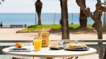 Ein Tisch mit Frühstücksangeboten, einem Getränk und einer Tasche mit QR-Code, mit Blick auf einen Strand mit Palmen und dem Meer.
