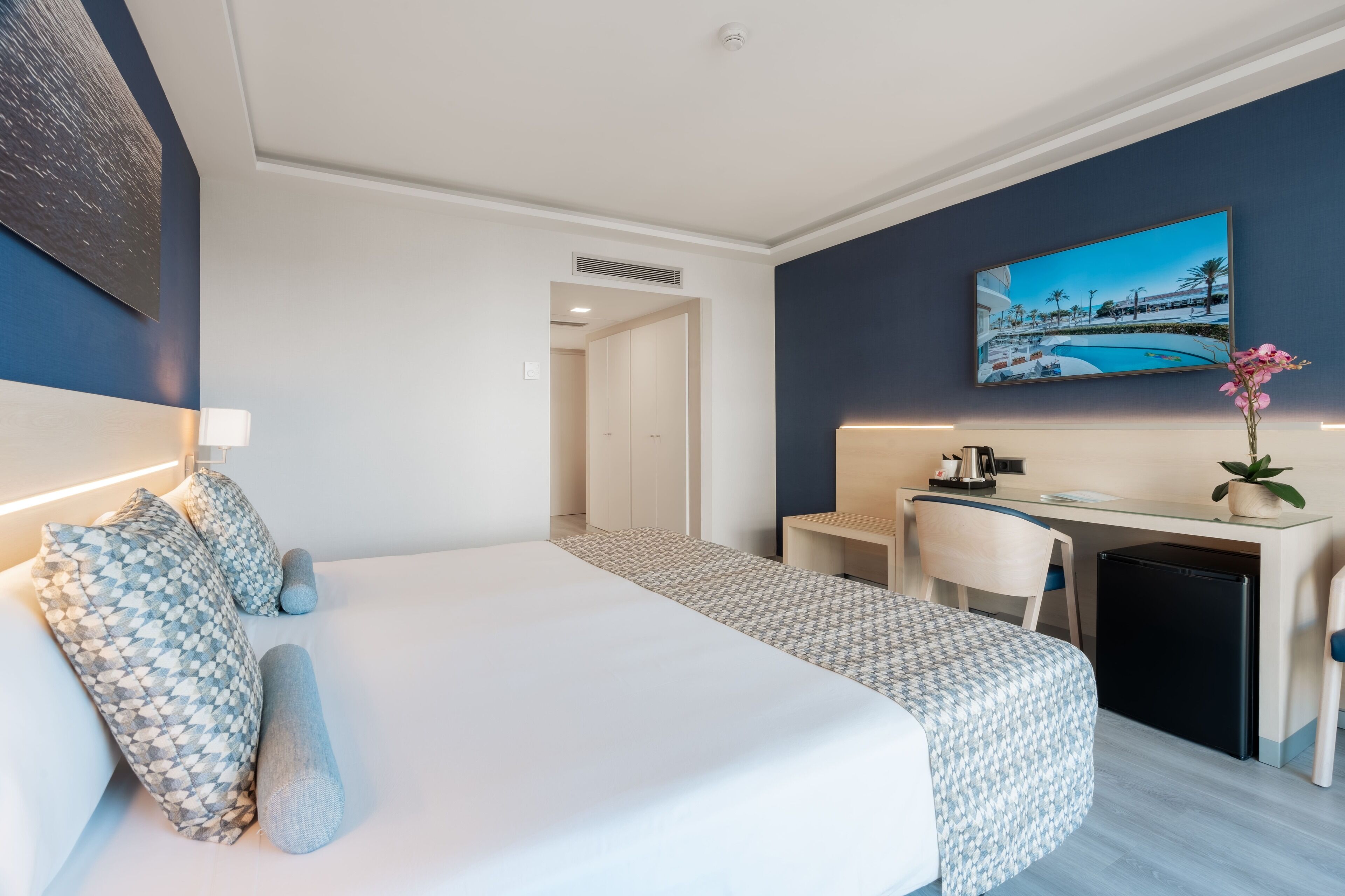 A hotel room with a bed, desk, chair, and a small refrigerator.