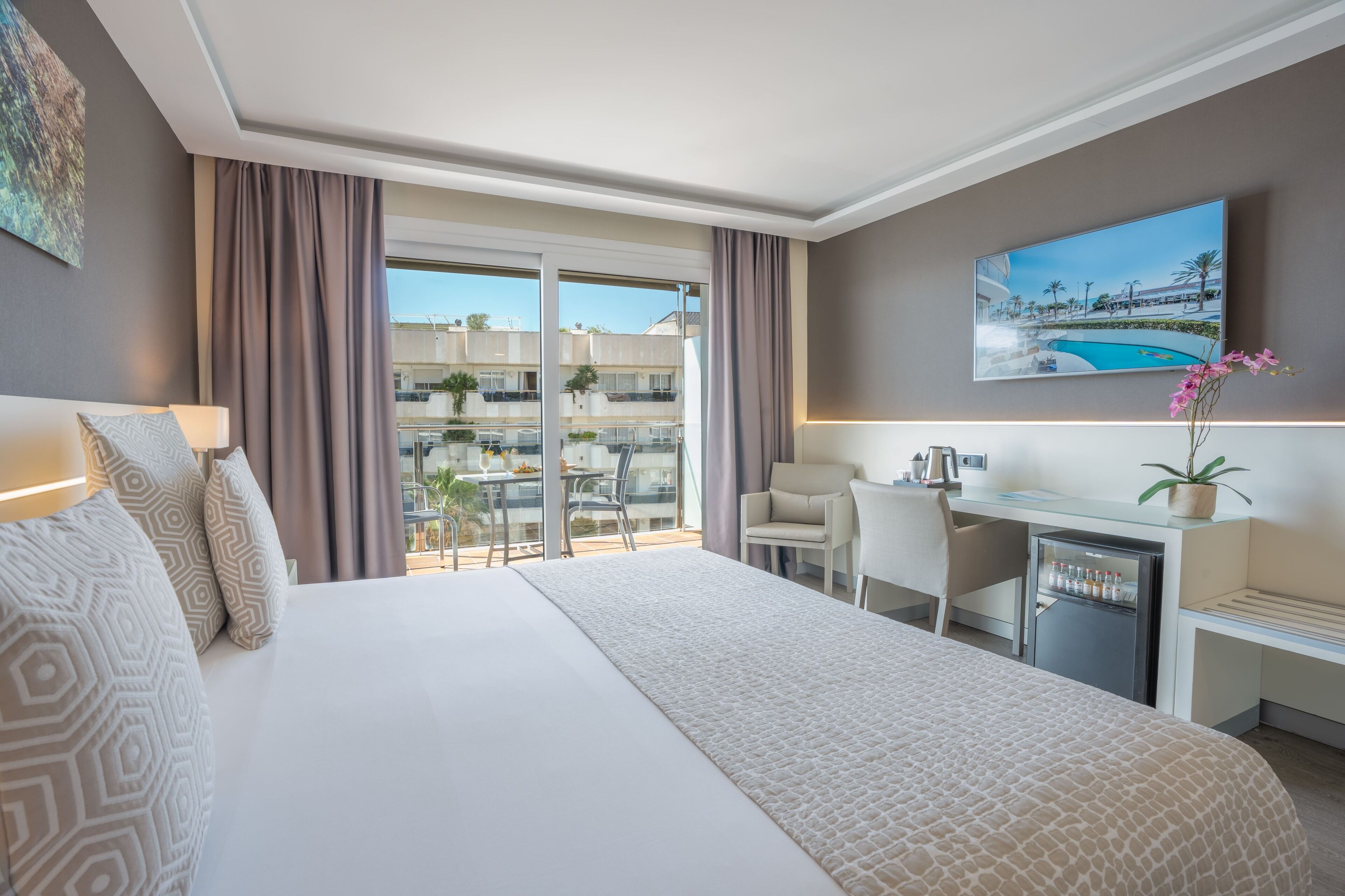 Una camera d'albergo con un letto grande, una scrivania, una sedia, un piccolo frigorifero e un balcone con vista su edifici e una piscina.
