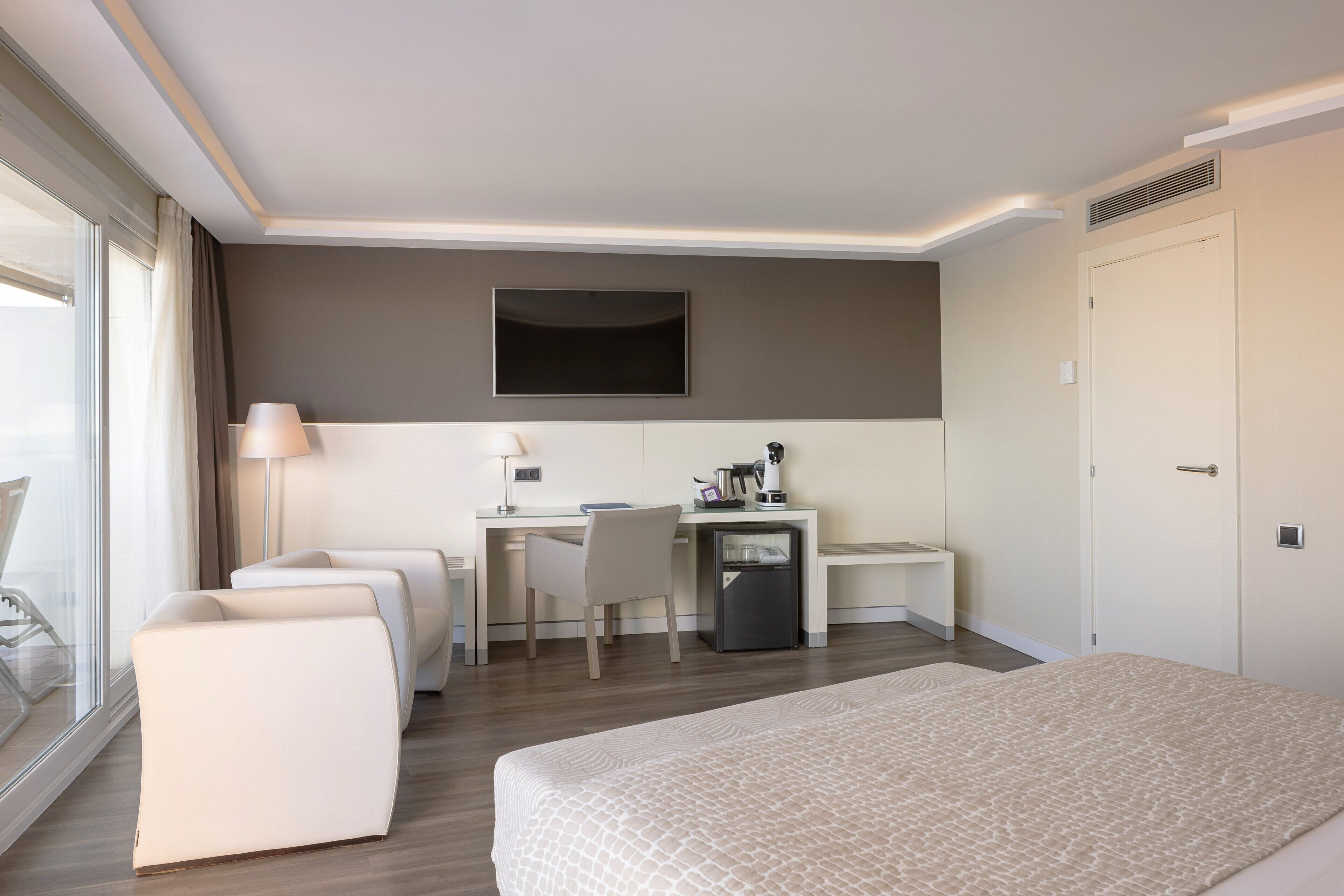 Una habitación de hotel moderna con una cama grande, un escritorio con silla, un refrigerador pequeño y un televisor montado en la pared.