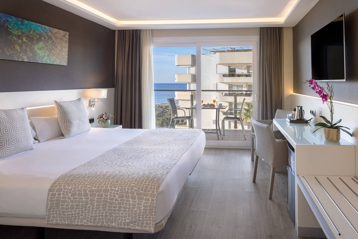 A modern hotel room with a large bed, a desk, and a balcony with a view of the sea.
