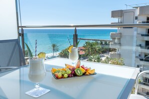 A balcony with a table featuring a fruit platter and two drinks, overlooking a coastal view.