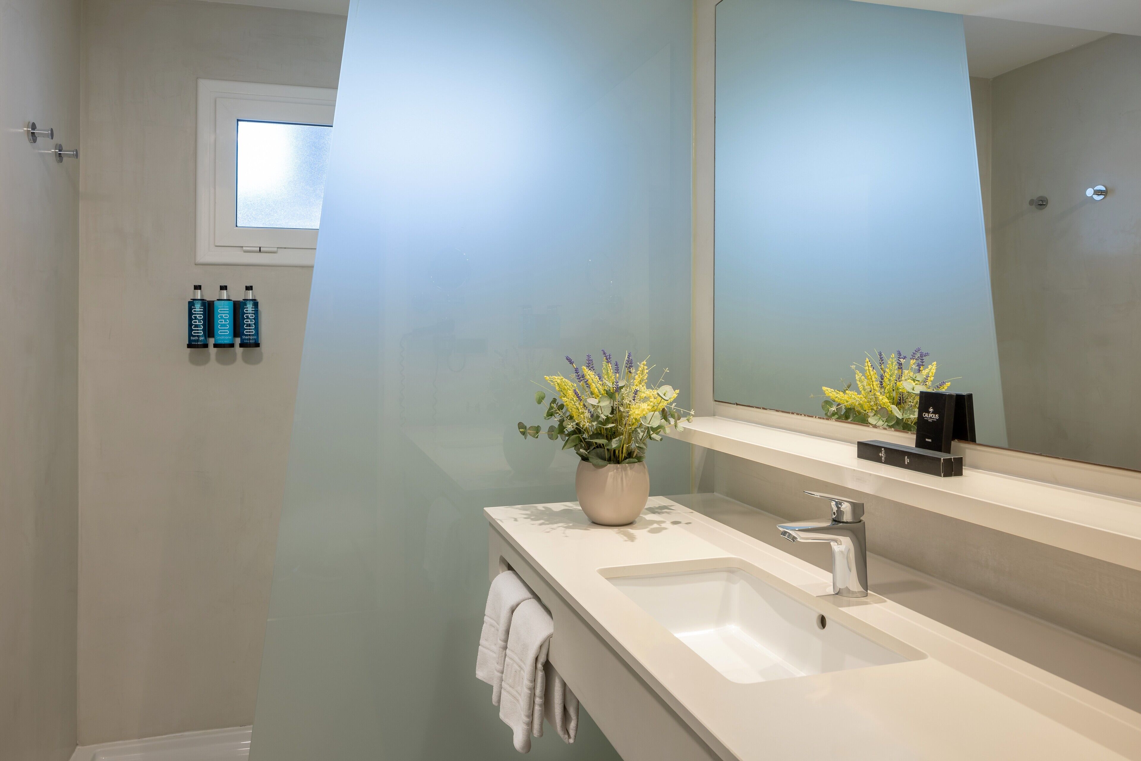 A modern bathroom with a large mirror, a sink, and a vase of flowers. There are three bottles on the wall and a towel hanging on the side.