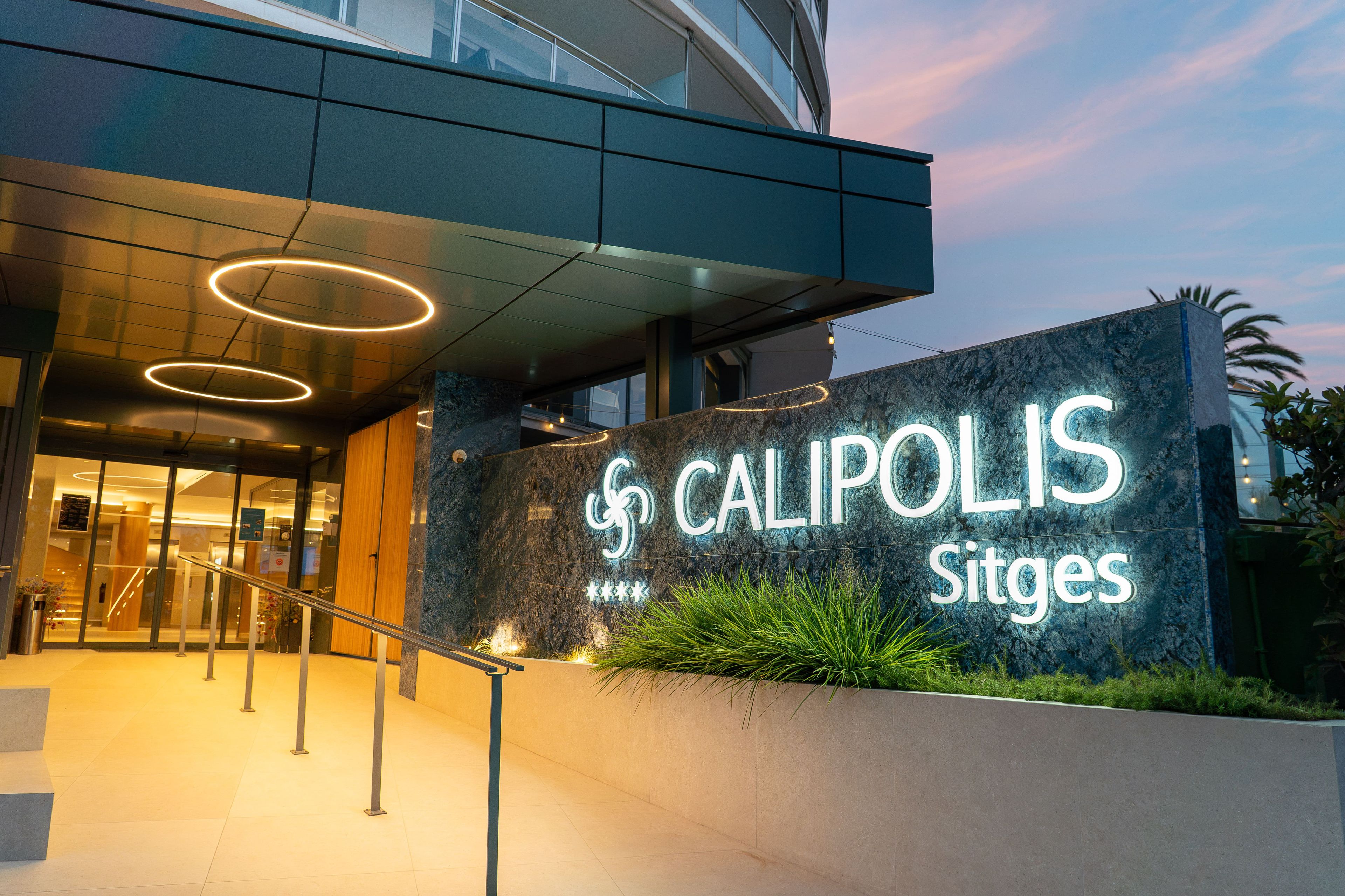 Entrada de un hotel con un letrero que dice 'CALIPOLIS Sitges' y un diseño arquitectónico moderno.