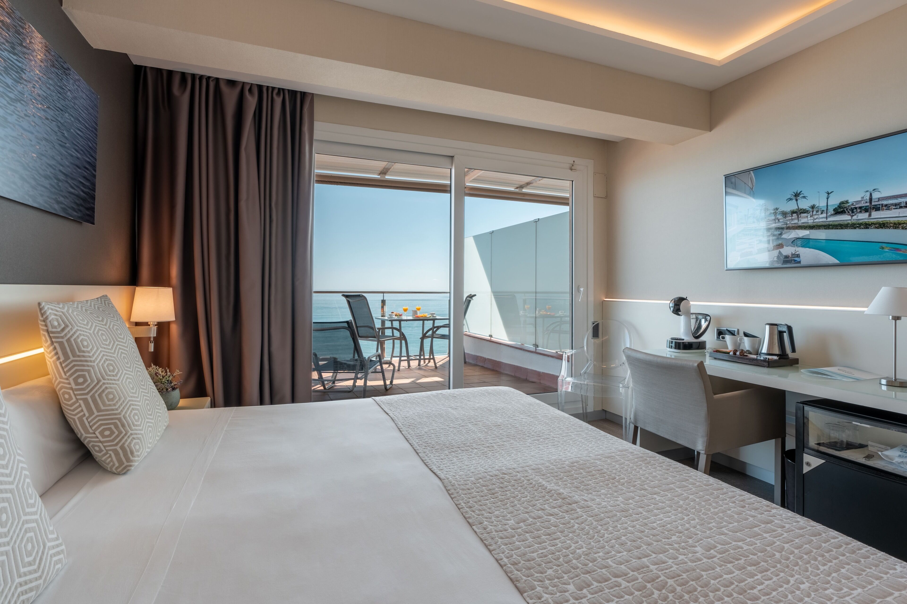 Habitación de hotel con una cama grande, un escritorio, una silla, un balcón con vistas al mar y un cuadro en la pared.
