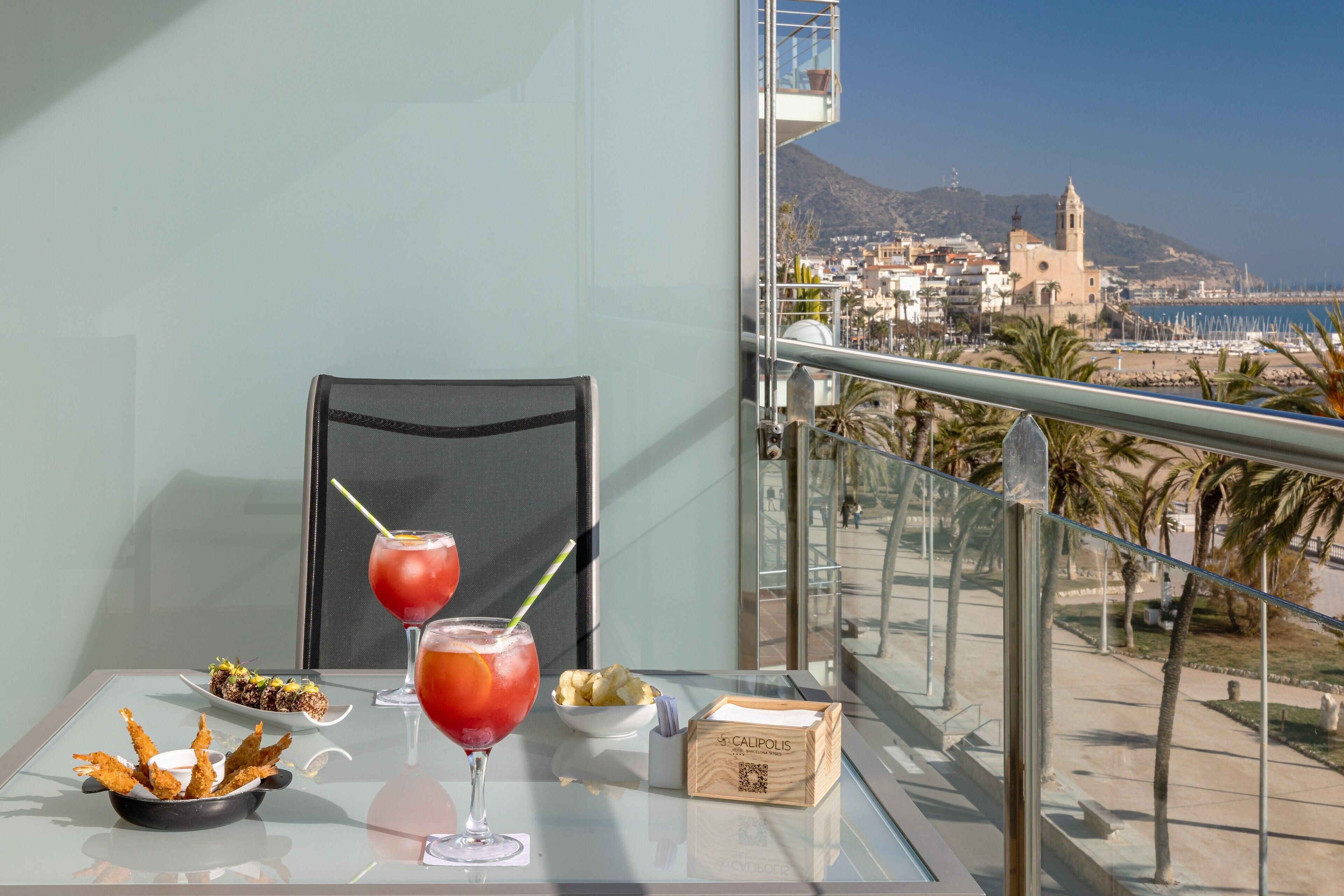A balcony with a table set with food and drinks, a chair, and a view of a coastal town with buildings and a mountain in the background.