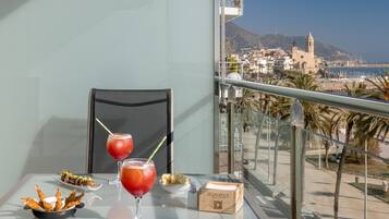 A balcony with a table set with food and drinks, a chair, and a view of a coastal town with buildings and a mountain in the background.