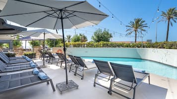 A poolside area with lounge chairs and umbrellas, a clear blue pool, and palm trees in the background.
