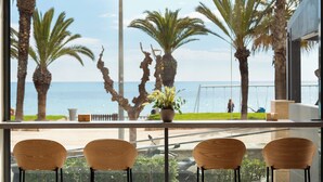 A view from a window with a potted plant, three wooden chairs, and a view of the sea and palm trees. - Hotel Calipolis (Sitges)