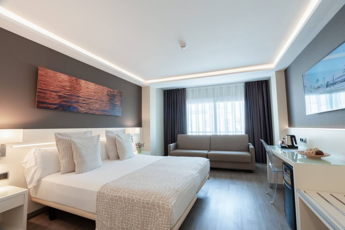 A modern hotel room with a bed, a sofa, a desk, and a dining area.