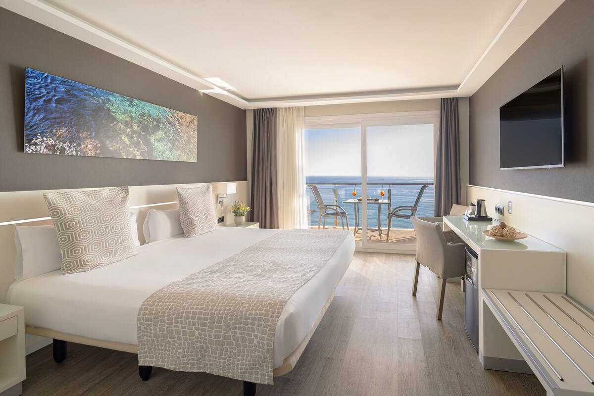 A modern hotel room with a large bed, a desk, and a balcony with a view of the sea.