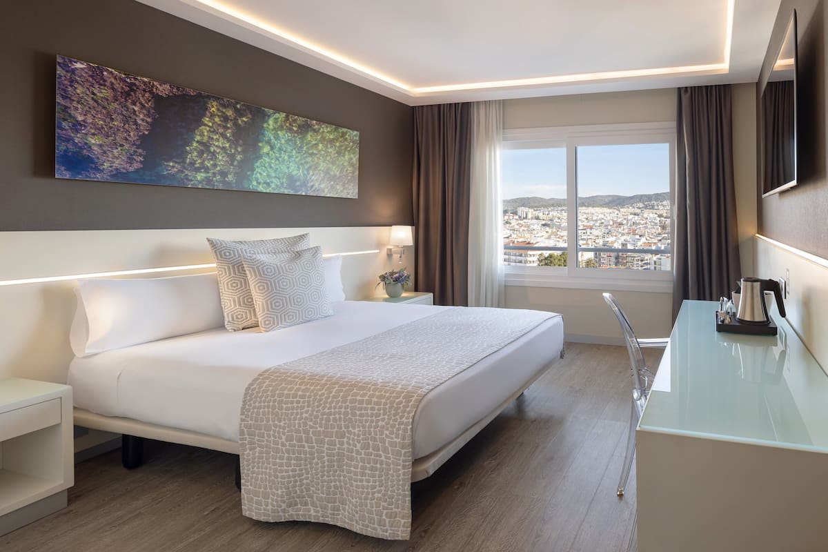 A modern hotel room with a large bed, bedside tables, a desk, and a city view.