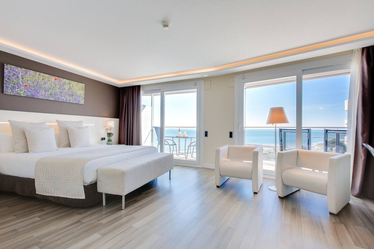 A modern hotel room with a large bed, a sofa, and a balcony with a view of the sea.