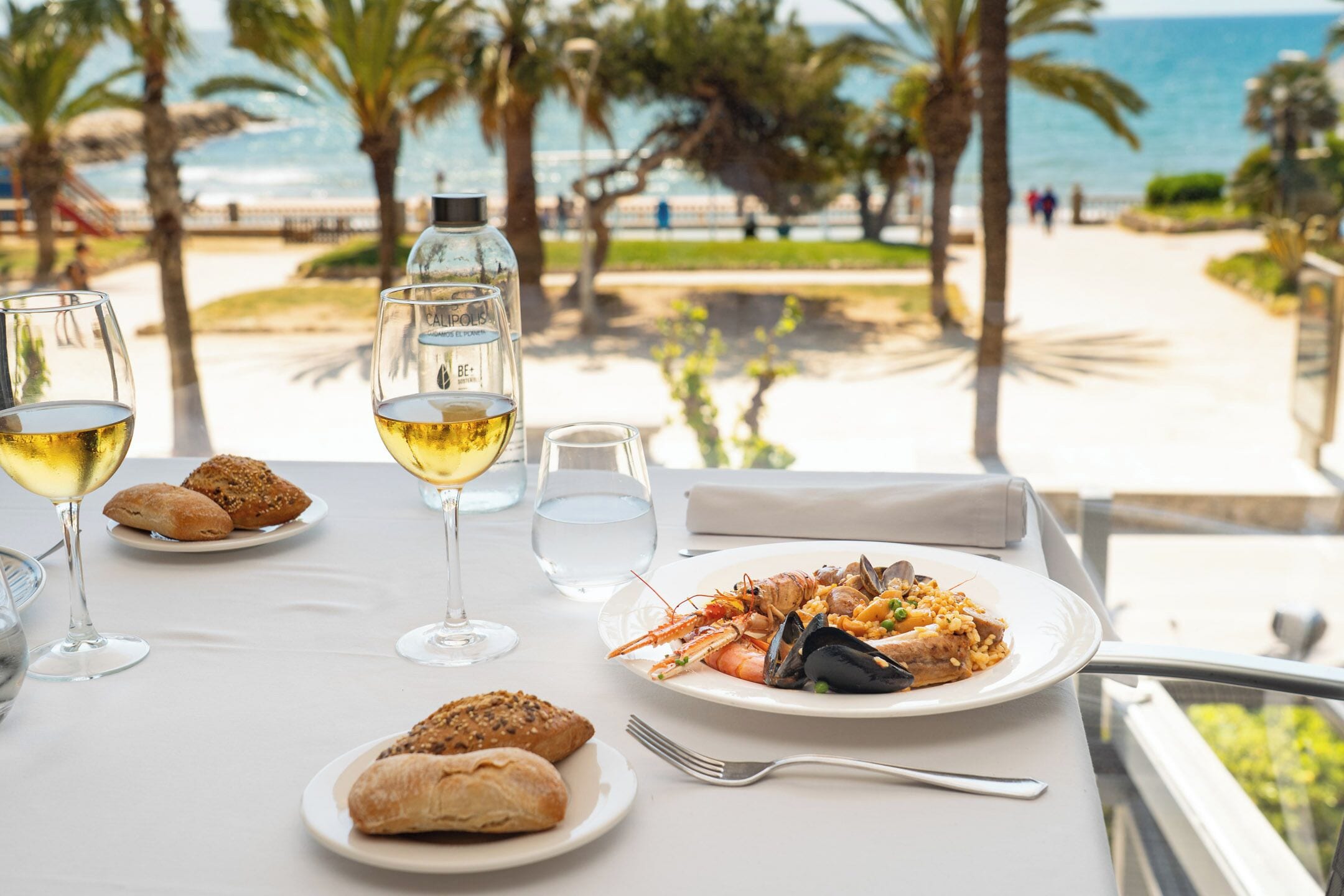 A table with a seafood dish, wine glasses, and bread, overlooking a beach and palm trees.