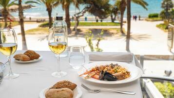 Una mesa con un plato de mariscos, copas de vino y pan, con vista a una playa y palmeras.