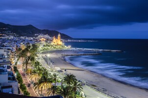 A coastal cityscape with a prominent church, a beach, and a promenade lined with palm trees.