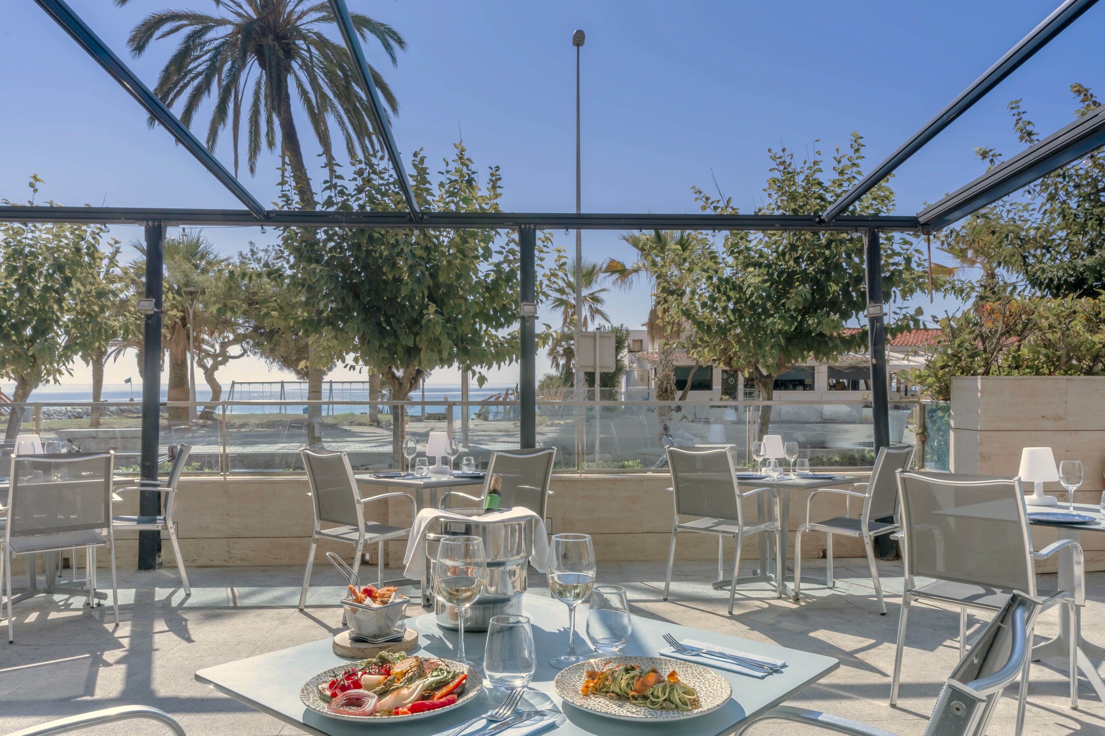 Un espacio de comedor al aire libre con mesas y sillas, con vistas a un paisaje escénico con palmeras y una masa de agua.
