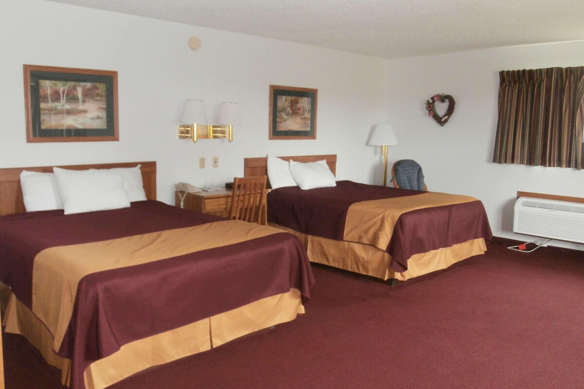 A hotel room with two beds, a desk, a chair, a lamp, and a wall-mounted heater.
