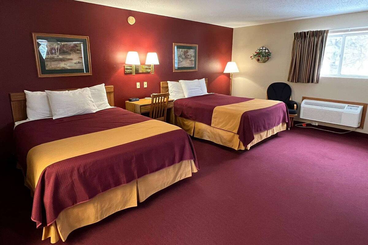 A hotel room with two beds, a desk, a chair, a window with curtains, and wall-mounted lamps.