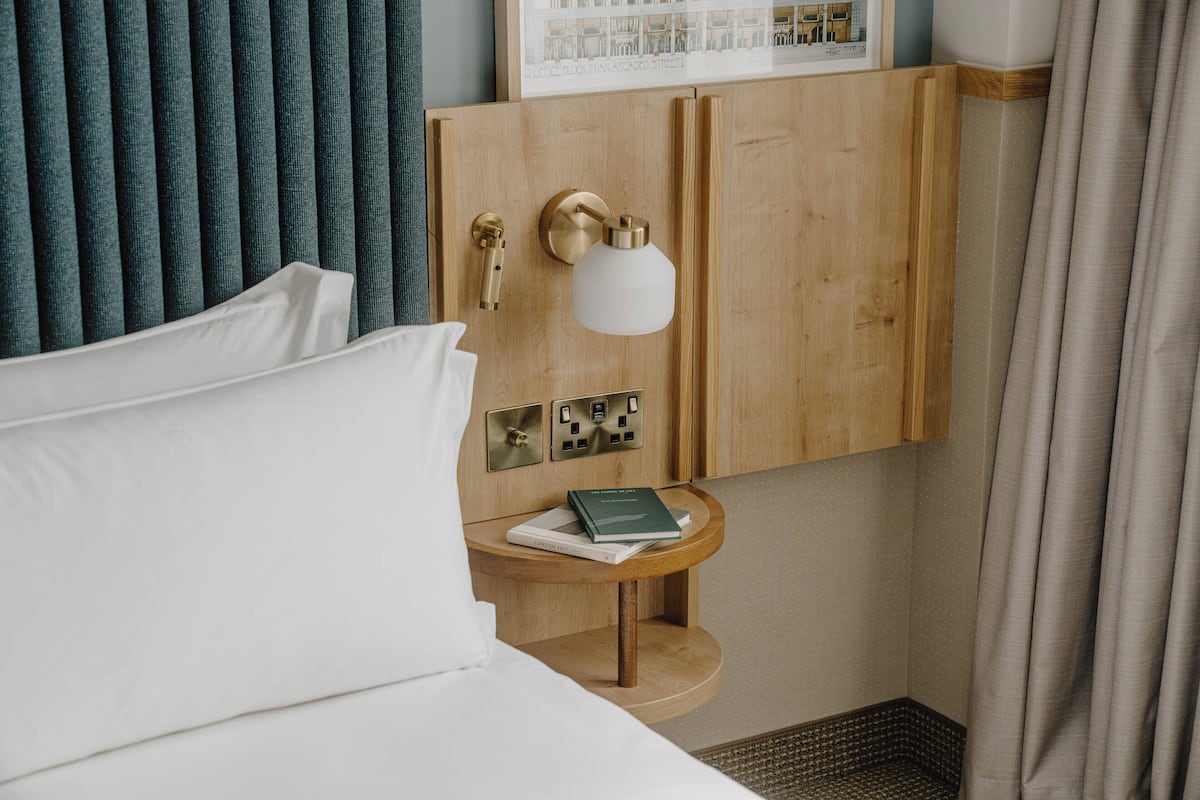 A hotel room with a bed, a wooden nightstand with a book, a wall-mounted light, and a framed picture above the nightstand.