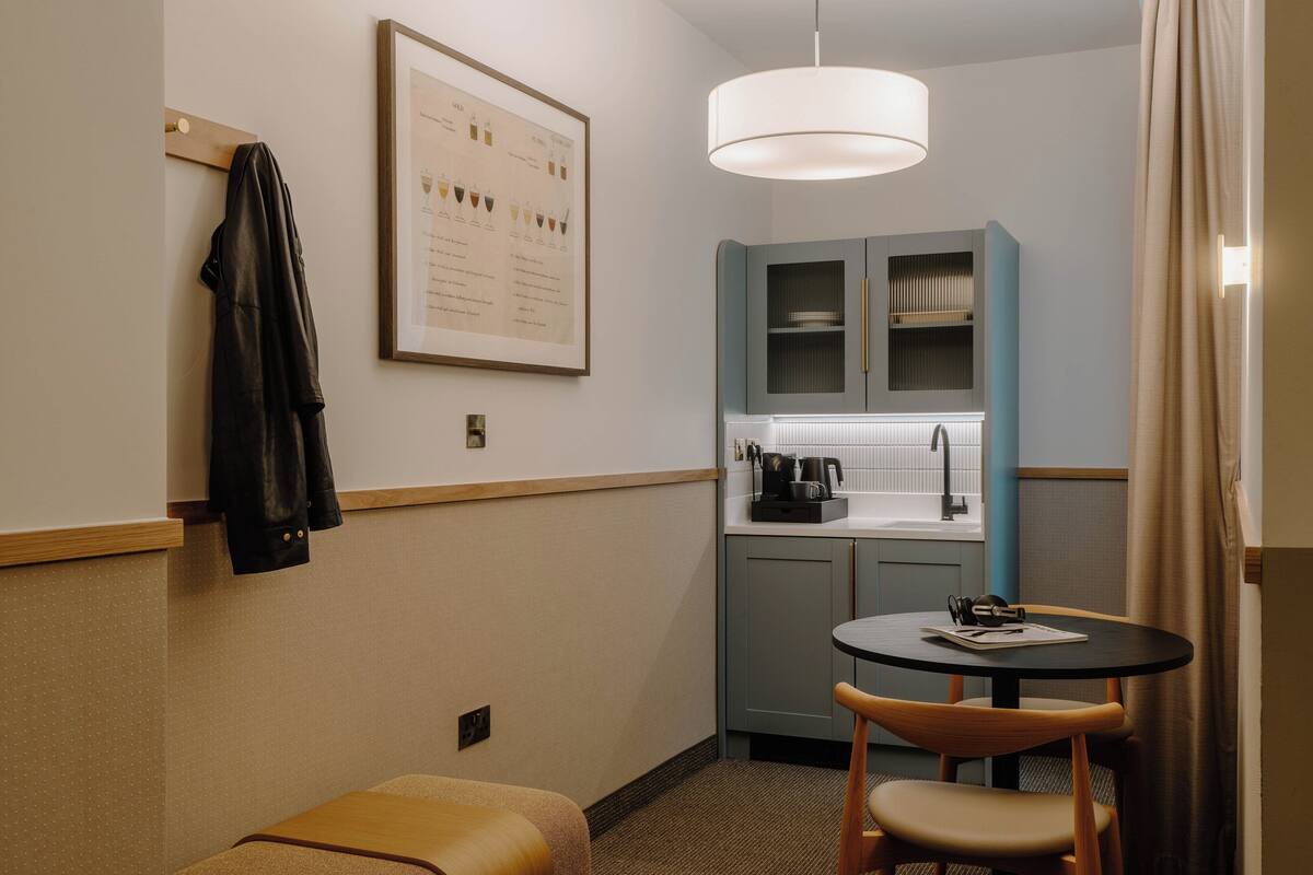 A compact hotel room with a kitchenette, a small dining table, a single chair, a coat rack, and a framed menu on the wall.