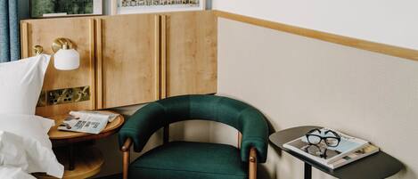 A hotel room with a green upholstered chair, a wooden cabinet, a small round table with a magazine and glasses, and a bed with white linens.