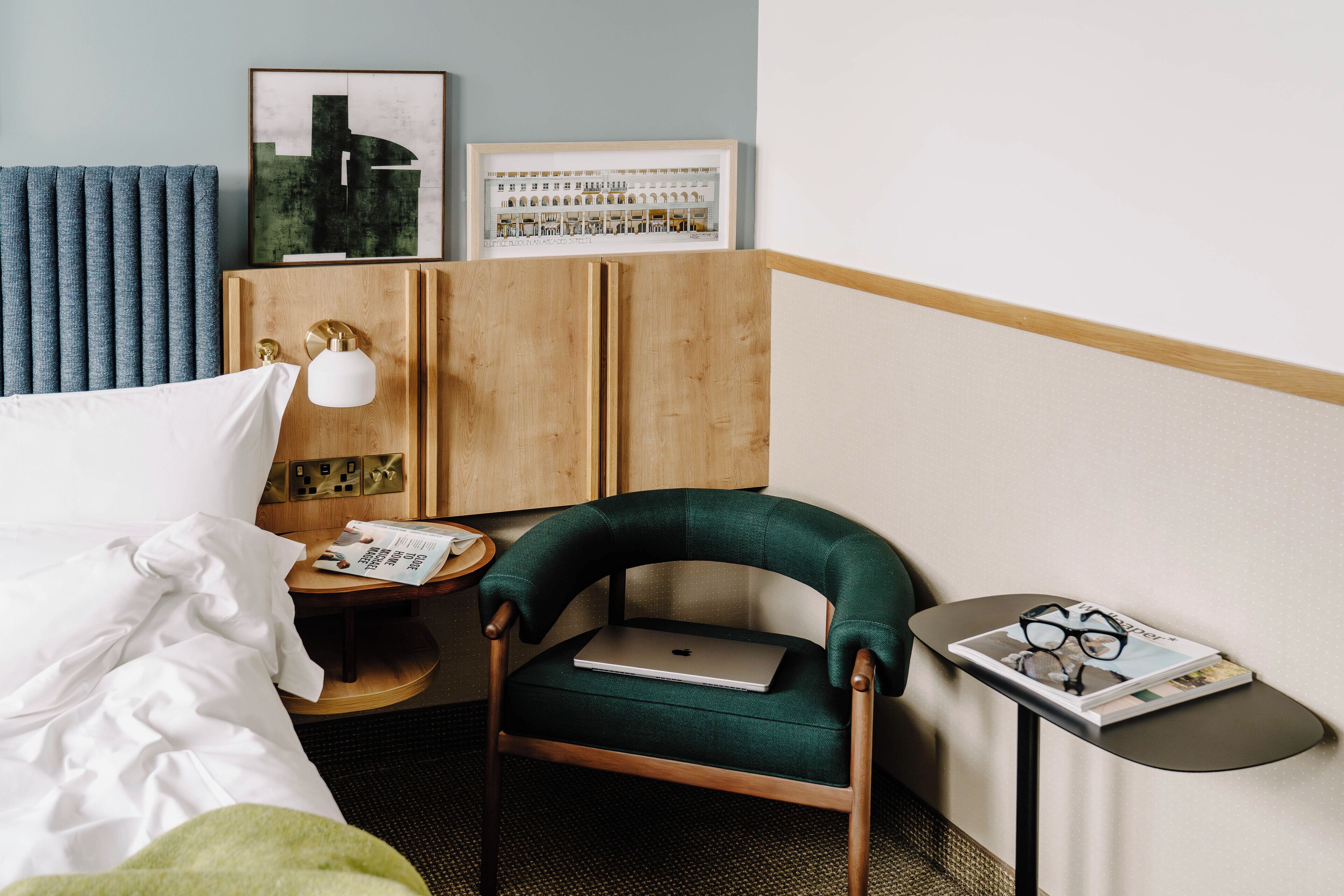 Ein Hotelzimmer mit Bett, Schreibtisch mit Laptop, Stuhl, Nachttisch mit Lampe und einer Wand mit gerahmten Bildern.