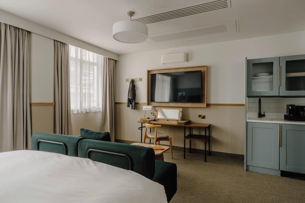 A hotel room with a large bed, a desk with a chair, a kitchenette, and a large window with curtains.