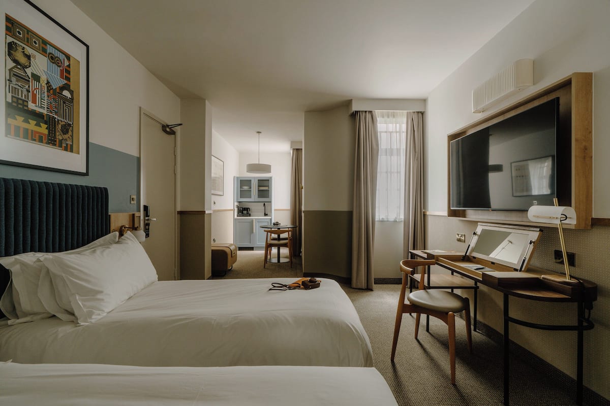 A hotel room with two beds, a desk with a chair, a television, and a kitchenette in the background.