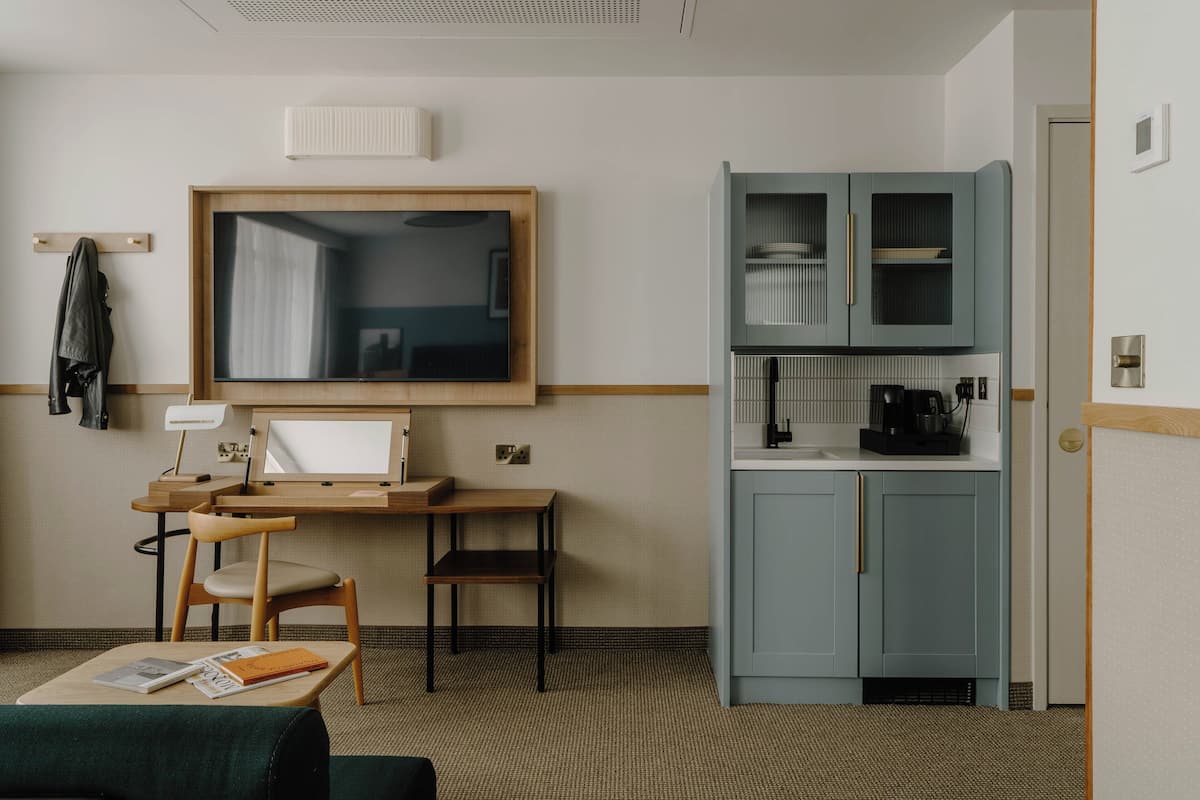 A hotel room with a wooden desk, a chair, a television, a kitchenette with cabinets, and a bed with a green blanket.