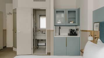 Habitación de hotel con cama, baño con lavamanos y espejo, y kitchenette con armarios y cafetera.