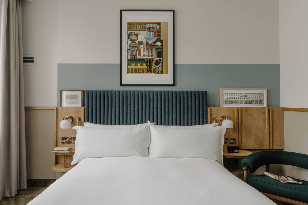 A hotel room with a large bed, a headboard, a framed picture on the wall, a nightstand with a lamp, and a chair.