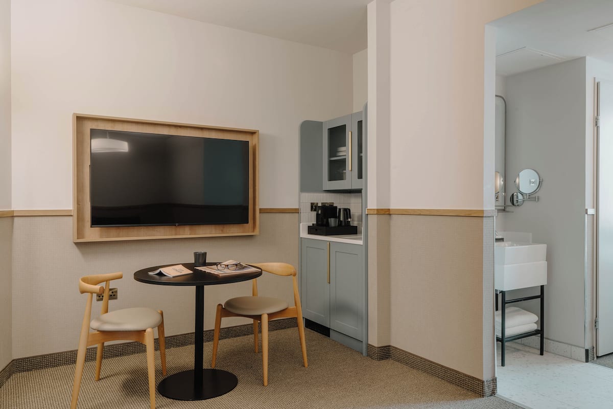 A hotel room with a TV, a small table with two chairs, a kitchenette, and a bathroom.