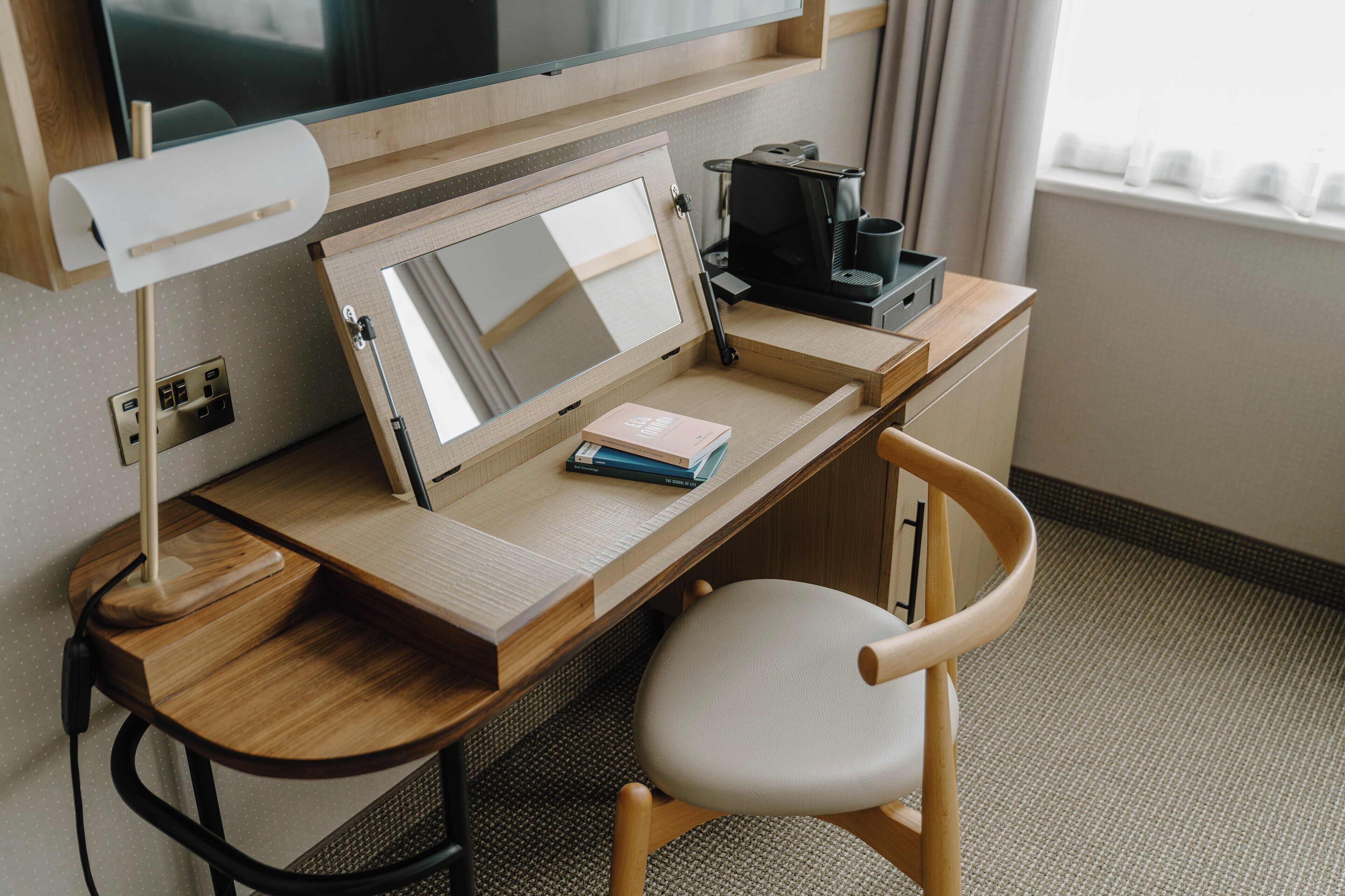 Un bureau en bois avec un écran d’ordinateur, une lampe, un clavier, une machine à café et une chaise.