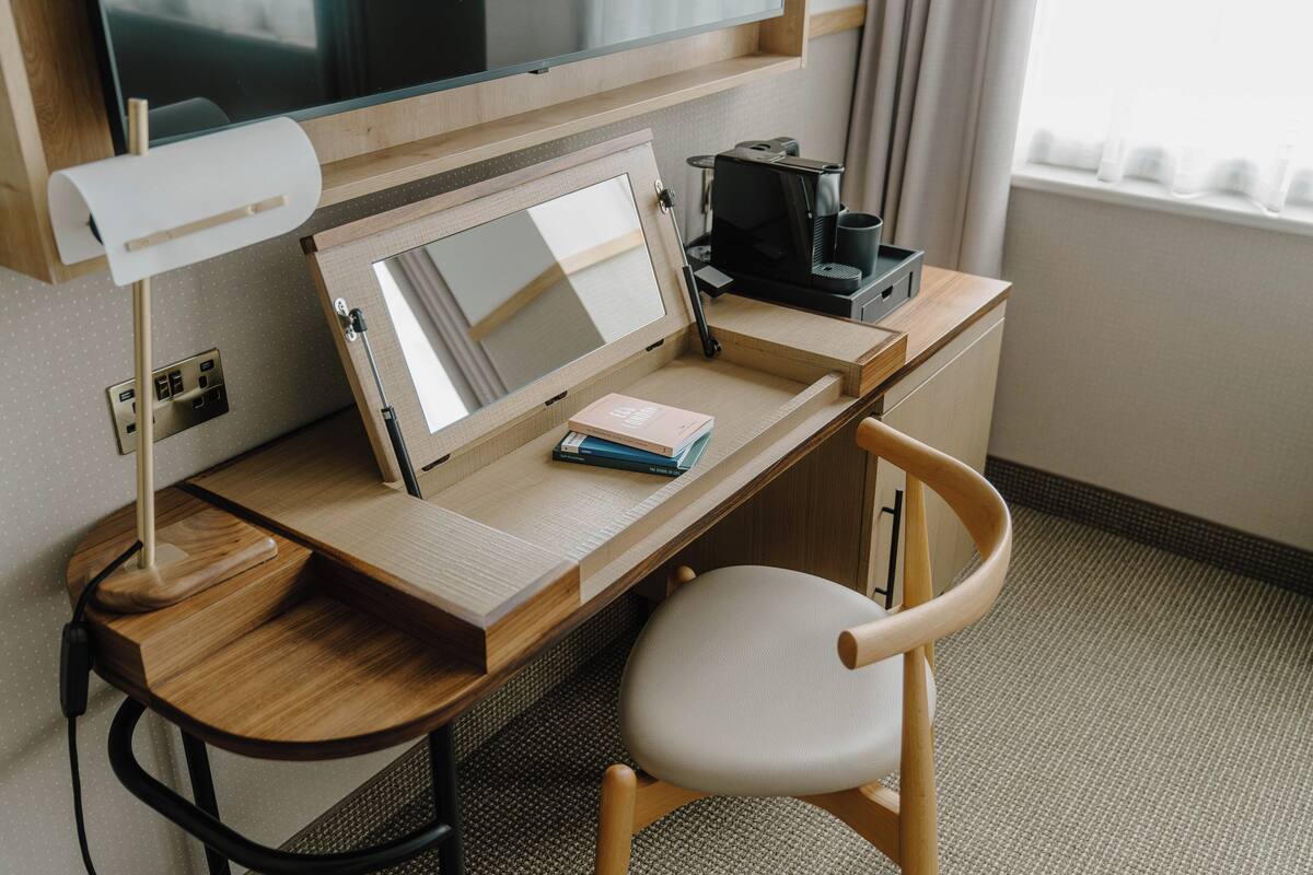 A wooden desk with a computer monitor, a lamp, a keyboard, a coffee maker, and a chair.