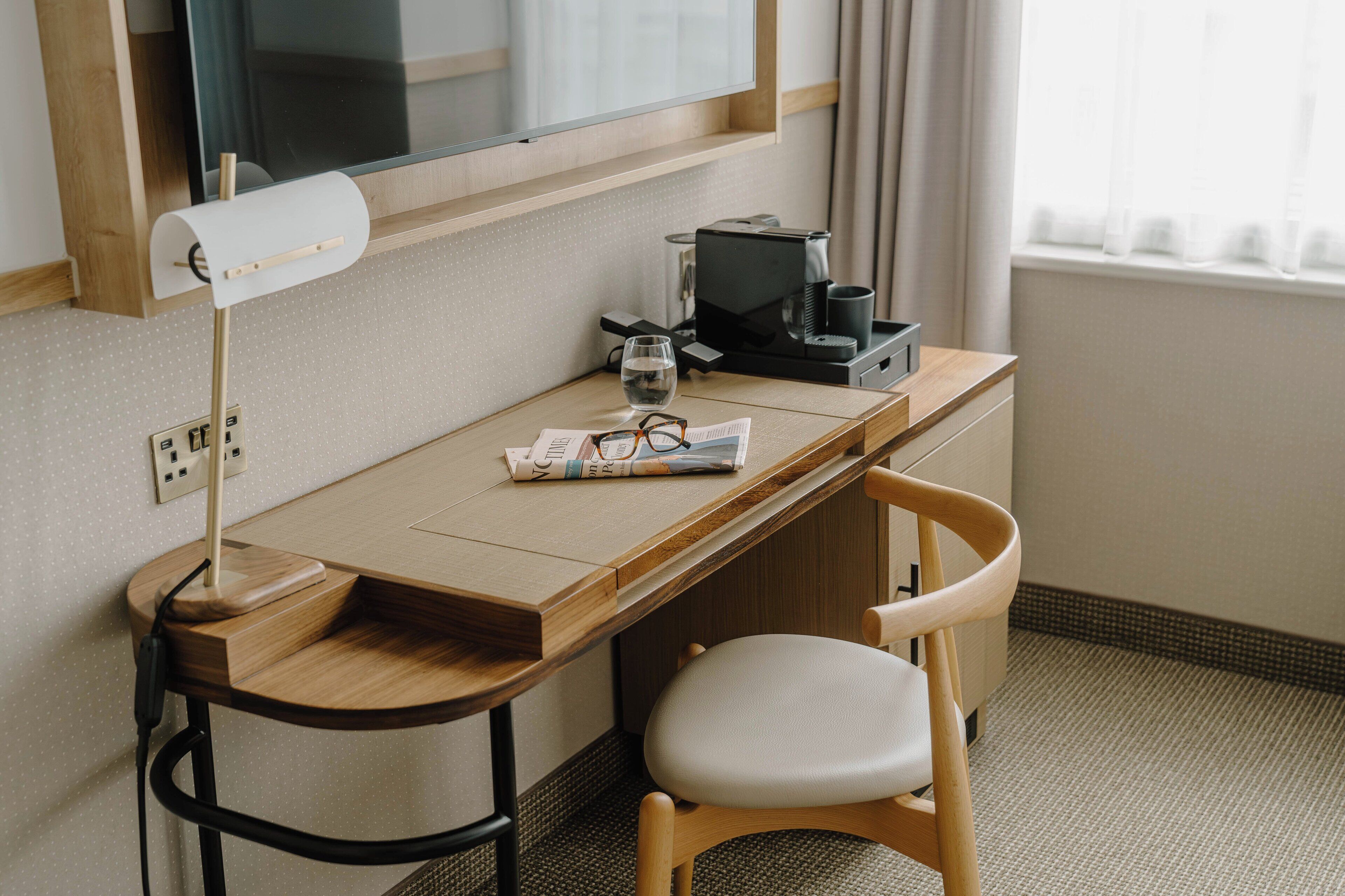 Habitación de hotel con un escritorio de madera, una silla, una lámpara de escritorio, una cafetera, un vaso de agua y una revista.