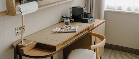 Habitación de hotel con un escritorio de madera, una silla, una lámpara de escritorio, una cafetera, un vaso de agua y una revista.