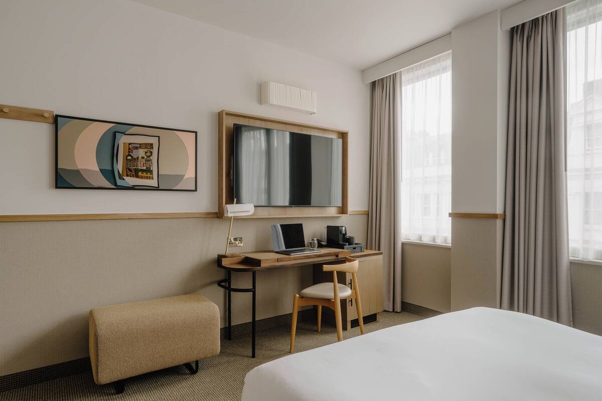 A hotel room with a bed, a desk with a laptop, a chair, a small ottoman, a mirror, and a window with curtains.