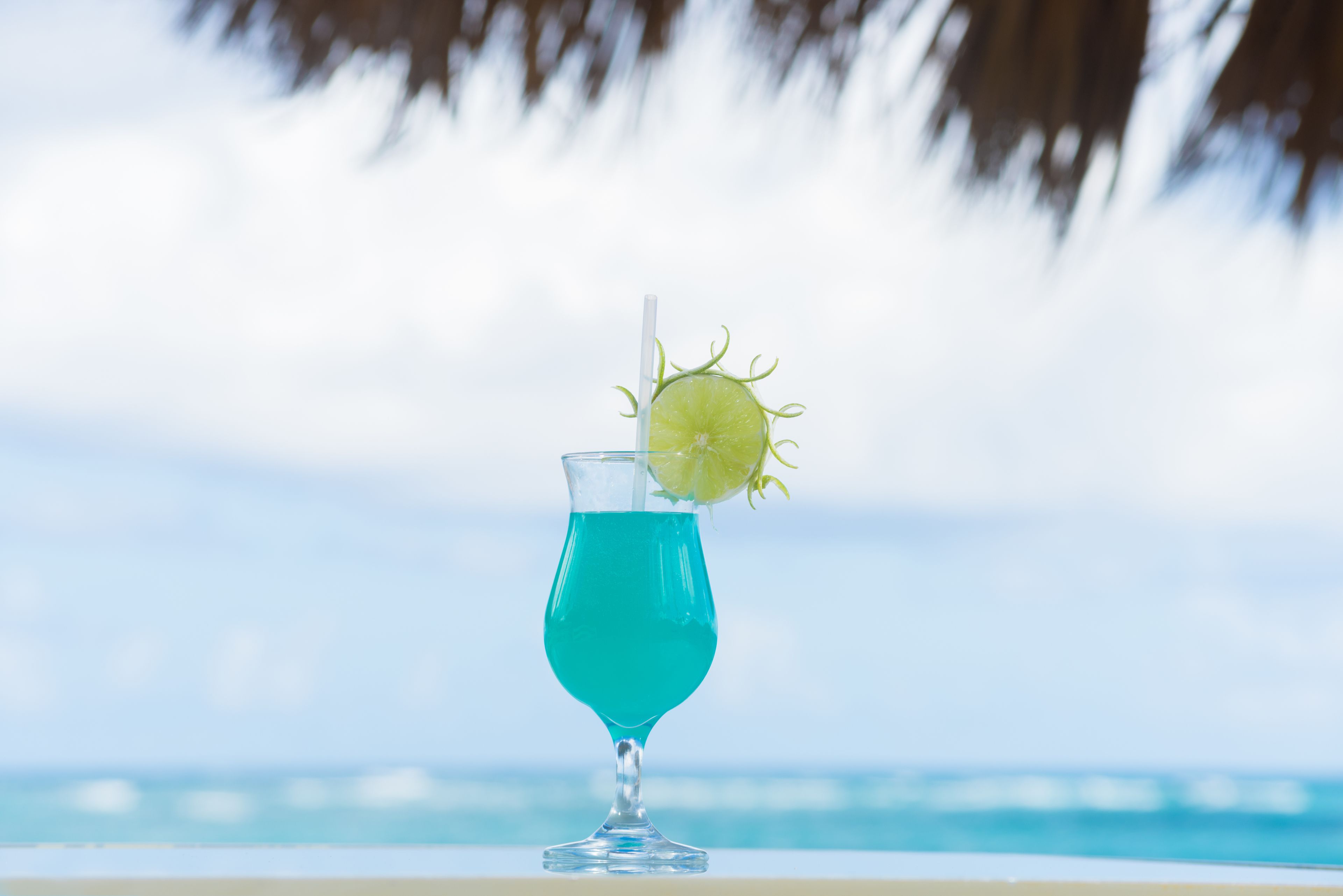 A clear glass with a lime garnish and a straw, placed against a blurred background of a beach and ocean.