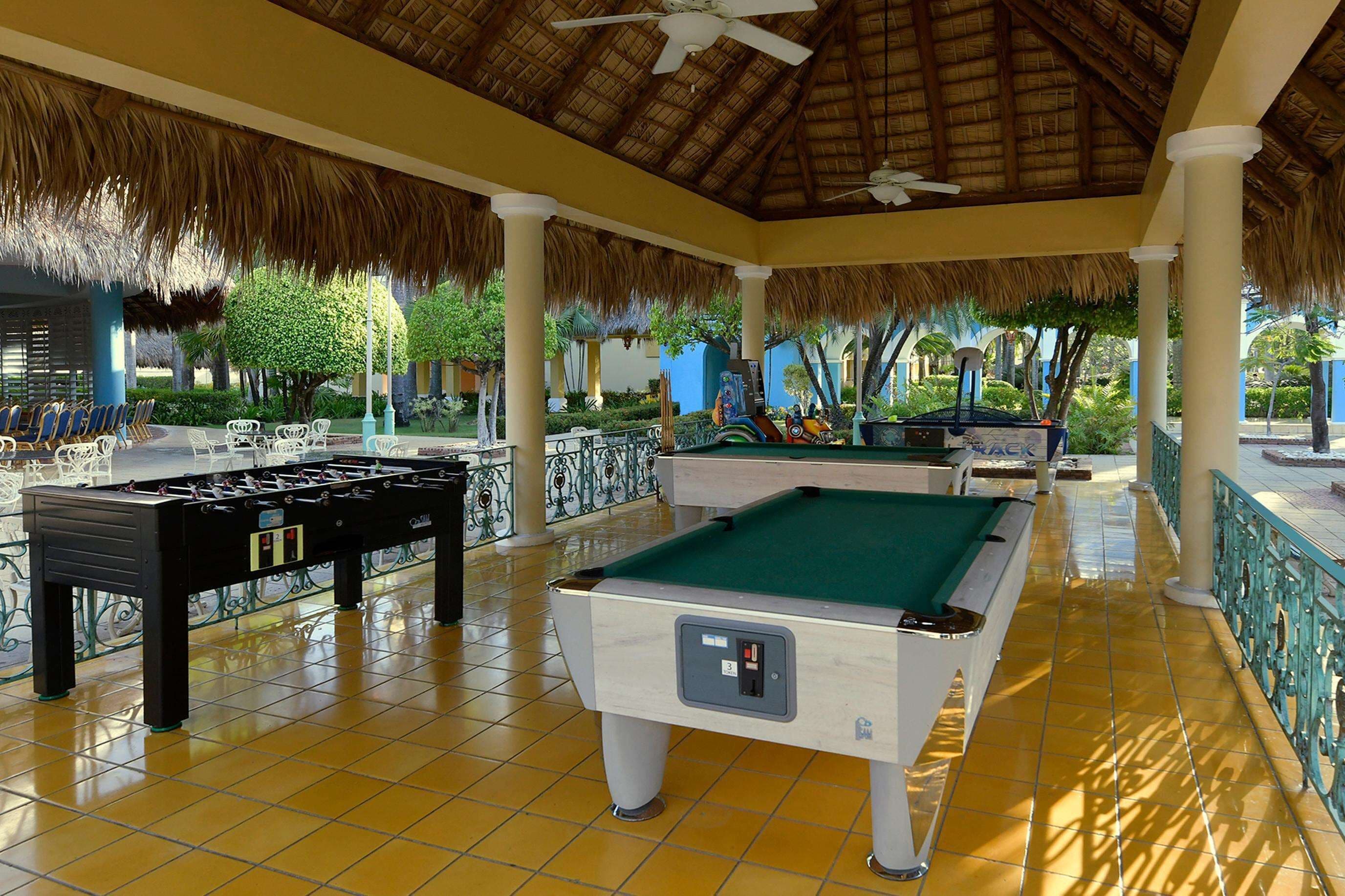 A pool table and foosball table in a covered outdoor area with tiled flooring and thatched roof.