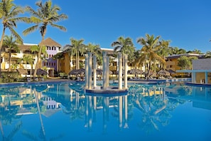 En pool vid ett semesterhotell, omgiven av palmer och byggnader med halmtak.
